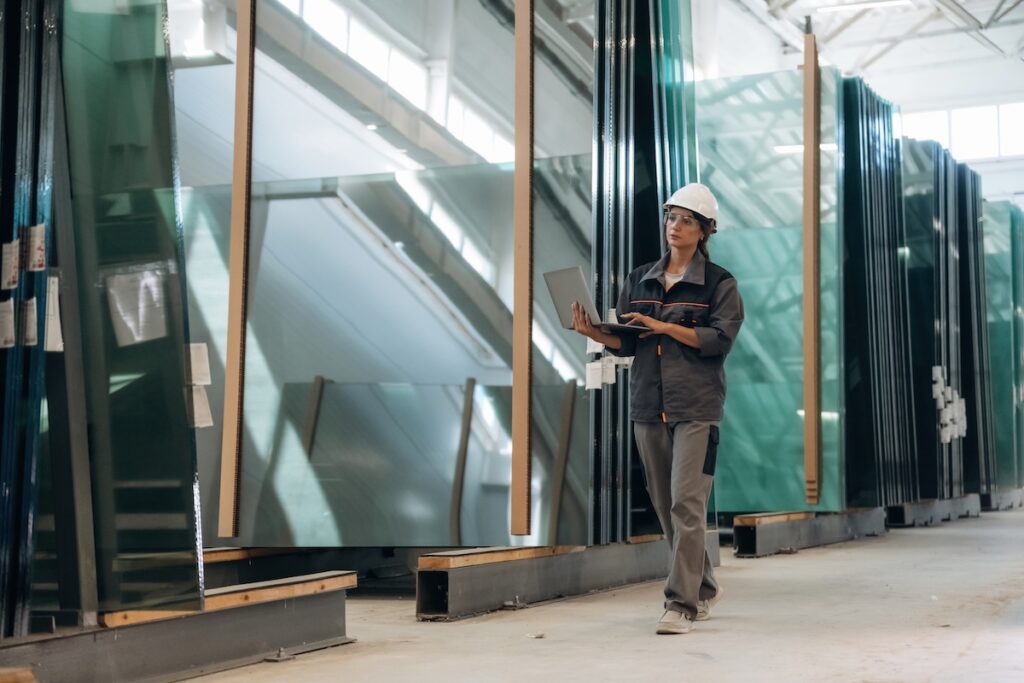 Factory worker inspecting large glass panels with a laptop in a warehouse